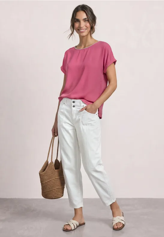Frau trägt pinke Bluse, weiße Jeans, Basttasche und weiße Sandalen vor hellem Hintergrund.