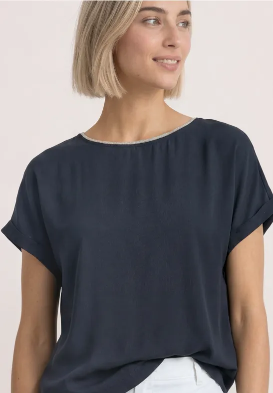 Eine junge Frau mit kurzen blonden Haaren trägt eine dunkelblaue, lockere Bluse vor hellem Hintergrund.