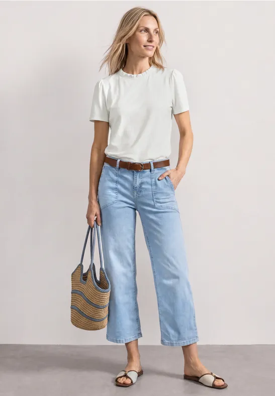 Frau in weißem T-Shirt und hellblauen Jeans, trägt gestreifte Tasche und flache Sandalen vor neutralem Hintergrund.