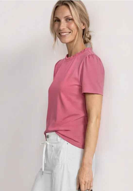 Zijaanzicht van een vrouw in een roze T-shirt en witte broek voor een lichte muur.