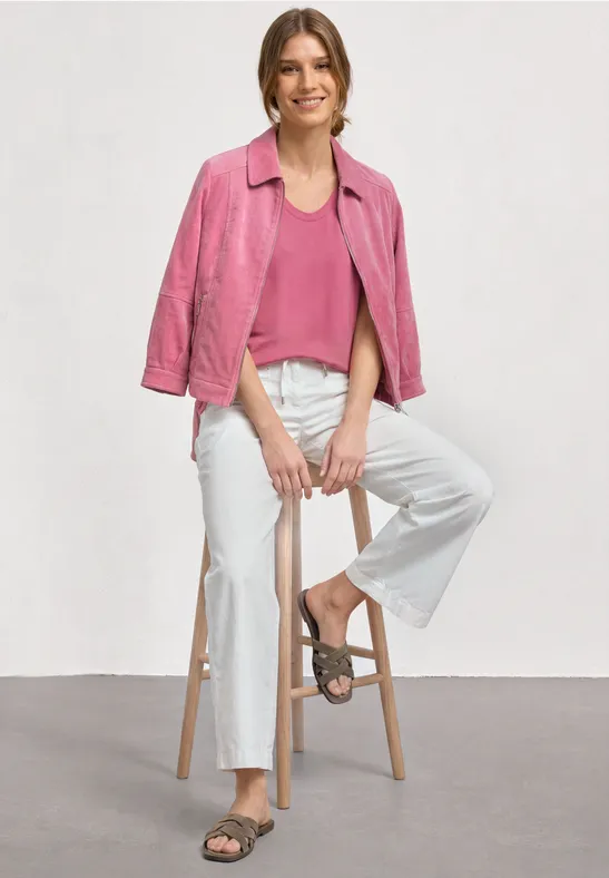 Personne assise sur un tabouret, porte une veste rose, un top rose, un pantalon blanc ample et des sandales.