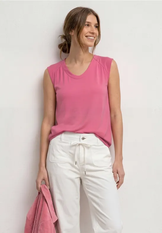 Femme portant un haut sans manches rose et un pantalon blanc, tenant un sac rose.