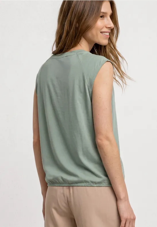 Une femme porte un débardeur vert sans manches et un pantalon beige, vue de côté, souriante.