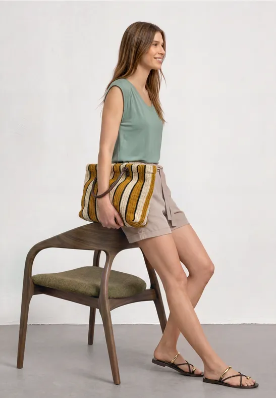 Une femme appuyée sur une chaise, en top vert, short beige et sandales, tenant un sac rayé.