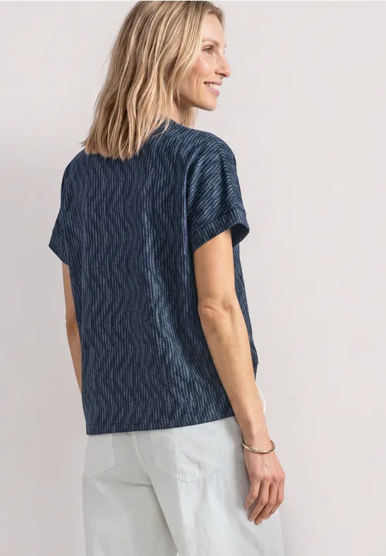 Femme de dos en tee-shirt bleu marine à motif ondulé et pantalon clair devant un fond neutre.