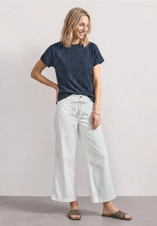 Femme debout devant un fond neutre, portant t-shirt bleu à rayures, pantalon large blanc et sandales.