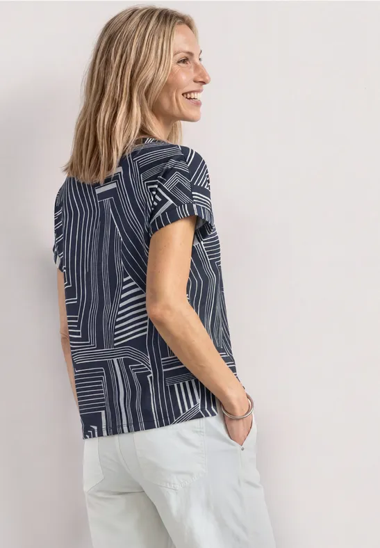 Femme vue de profil portant un t-shirt à motif bleu et pantalon blanc sur fond clair.