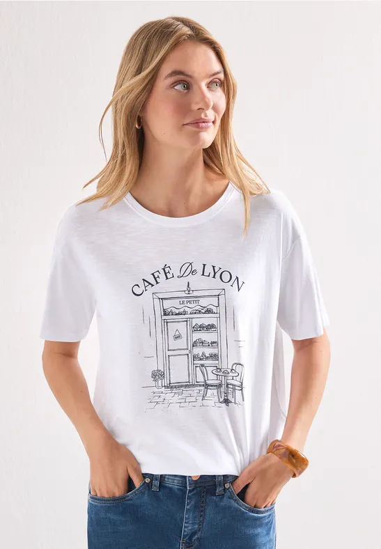 Frau in weißem T-Shirt mit Café-de-Lyon-Illustration, Jeanshose und locker fallenden Haaren vor hellem Hintergrund.