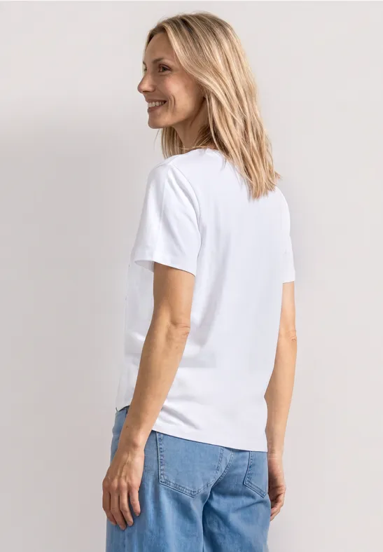 Eine blonde Frau steht seitlich, trägt ein weißes T-Shirt und blaue Jeans.