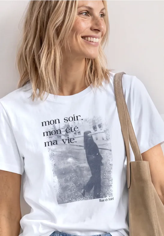 Eine Person trägt ein weißes T-Shirt mit dem Aufdruck „mon soir. mon été. ma vie.“, Katzenmotiv und eine beige Tasche.