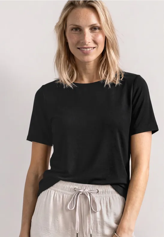 Frau in schwarzem T-Shirt und beiger Hose mit Kordelzug vor neutralem Hintergrund.