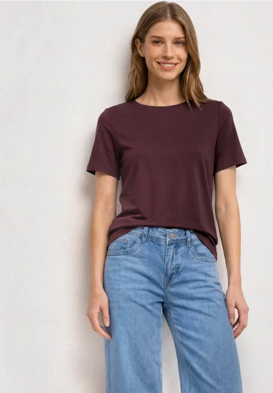 Een jonge vrouw in een paars t-shirt en blauwe jeans staat glimlachend voor een neutrale achtergrond.
