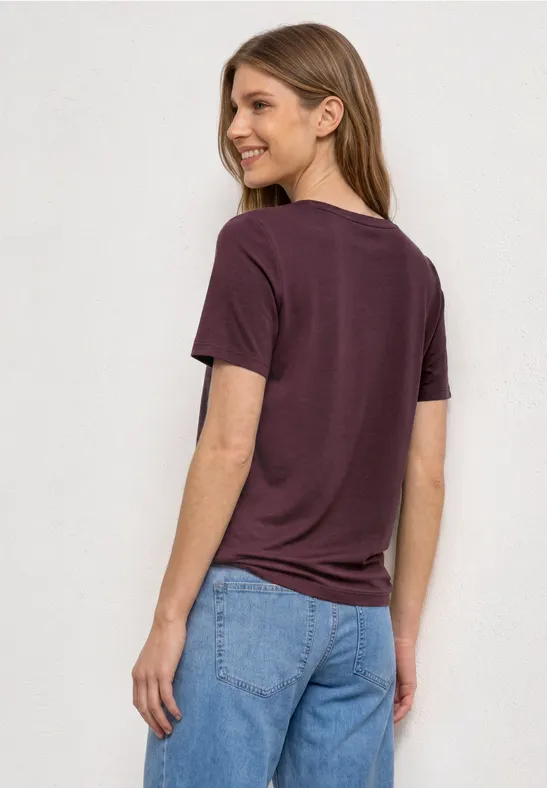 Vrouw van achteren in paars t-shirt en blauwe spijkerbroek tegen lichte achtergrond.
