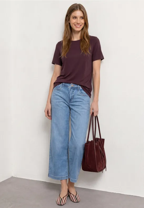 Vrouw leunt tegen witte muur, draagt paars shirt, wijde jeans, slippers en een rode tas.