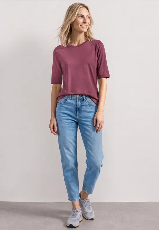 Une femme porte un t-shirt violet, un jean bleu et des baskets grises devant un mur uni.