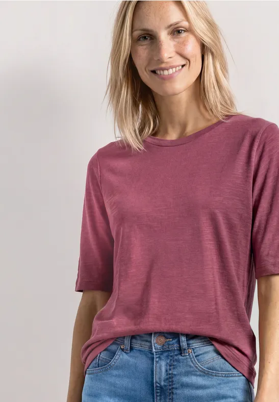 Femme blonde souriante en t-shirt rose à manches courtes, rentré dans un jean bleu, devant un fond clair.