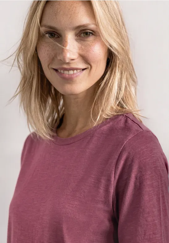 Personne au visage flouté, cheveux blonds mi-longs, portant un haut à manches longues rose.