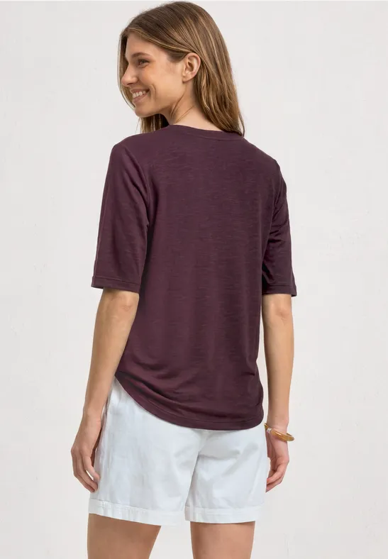 Vrouw van achteren met een losvallend donkerpaars shirt en witte korte broek voor een lichte achtergrond.