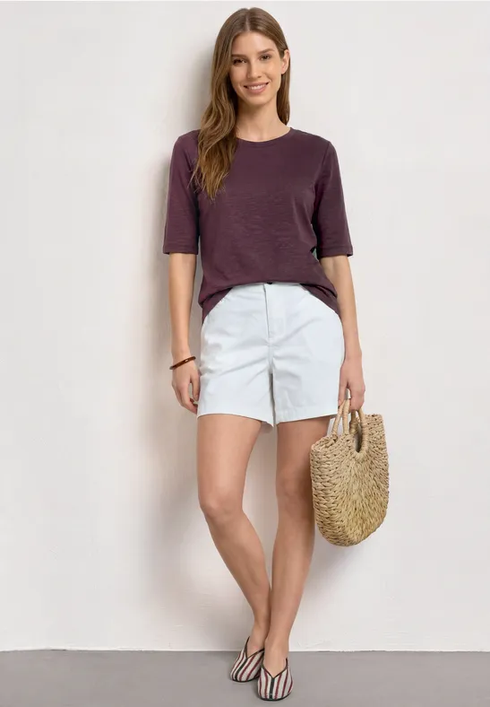 Vrouw in paarse top, witte korte broek en strooien tas tegen witte achtergrond.