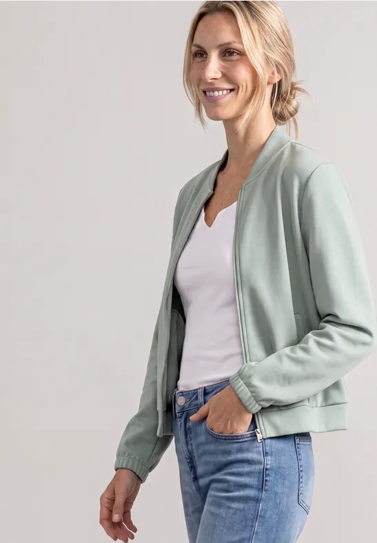 Femme souriante en bomber vert clair, top blanc et jean, posture décontractée
