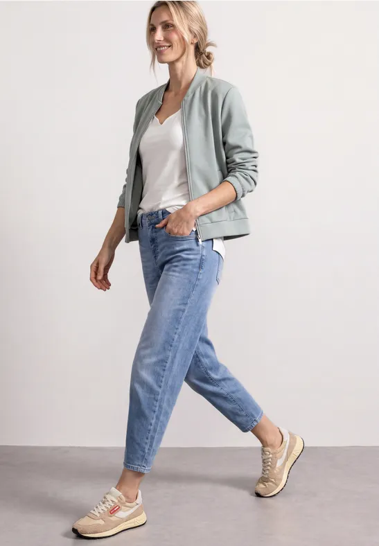 Femme en bomber gris, T-shirt blanc, jean bleu et baskets beiges marchant de façon décontractée.