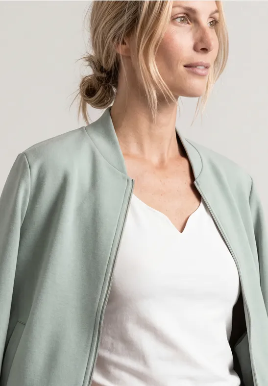 Femme blonde portant un t-shirt blanc et un blouson vert menthe, regard tourné de profil.