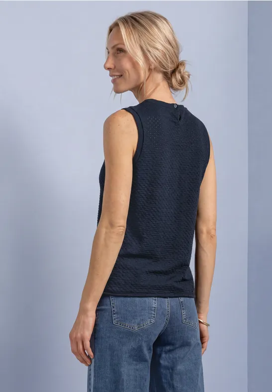 Frau im Profil, sie trägt ein ärmelloses, dunkelblau strukturiertes Top und Jeans vor hellem Hintergrund.