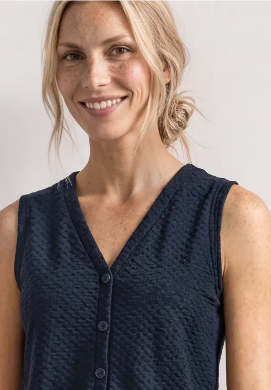 Lächelnde Frau mit blonden Haaren in dunkelblauer, ärmelloser V-Neck-Bluse mit Knöpfen.