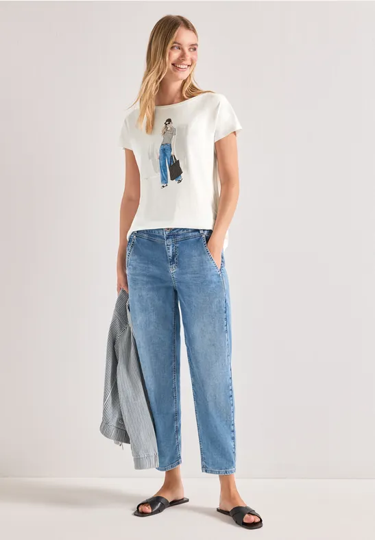 Frau in weißem T-Shirt, lockeren blauen Jeans und Sandalen hält eine graue Jacke.