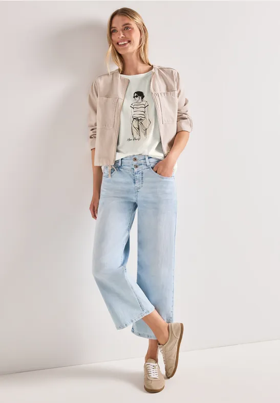 Vrouw in lichte jeans, wit grafisch t-shirt, beige blazer en loafers, staat nonchalant.
