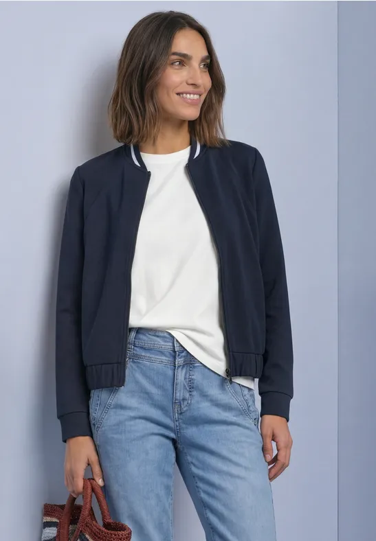 Frau in weißem T-Shirt, blauer Bomberjacke und Jeans, lächelt und trägt Tasche.