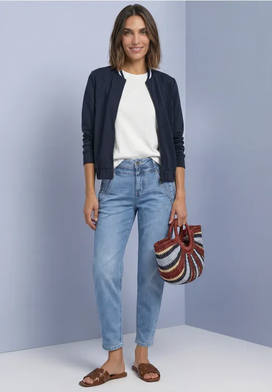 Eine Frau in weißem Shirt, dunkelblauer Strickjacke, Jeans und Sandalen trägt eine bunte Umhängetasche.