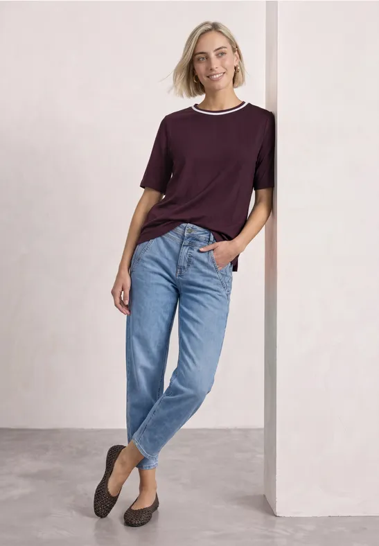 Eine lächelnde Frau lehnt entspannt an einer Wand, trägt blaues Jeans und lila T-Shirt.