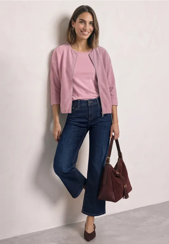 Une femme souriante appuyée contre un mur, en haut rose, jean bleu foncé et sac bordeaux.
