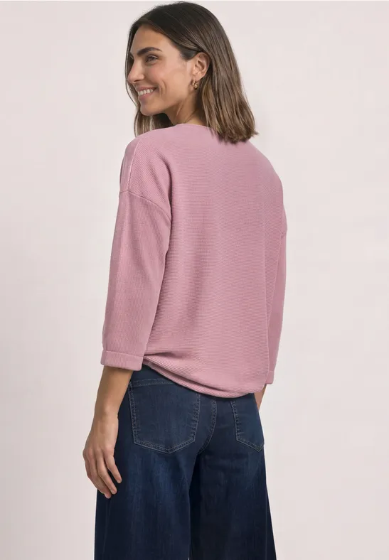 Une femme souriante en haut rose à manches 3/4 et pantalon large regarde par-dessus son épaule.