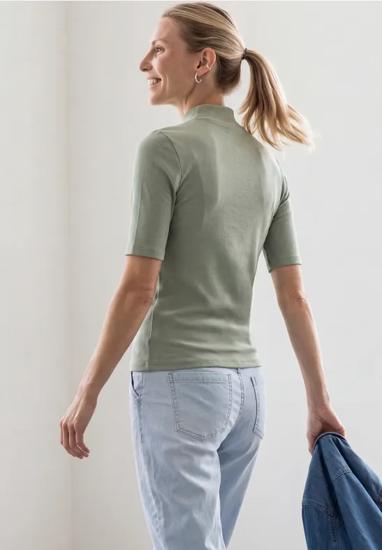 Frau von hinten in grünem Halbarmshirt und hellen Jeans, hält Jacke in der Hand.