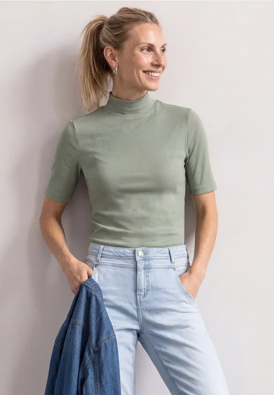 Eine lächelnde Frau in grünem Kurzarmshirt und hellblauen Jeans, die eine Jeansjacke vor hellem Hintergrund hält.
