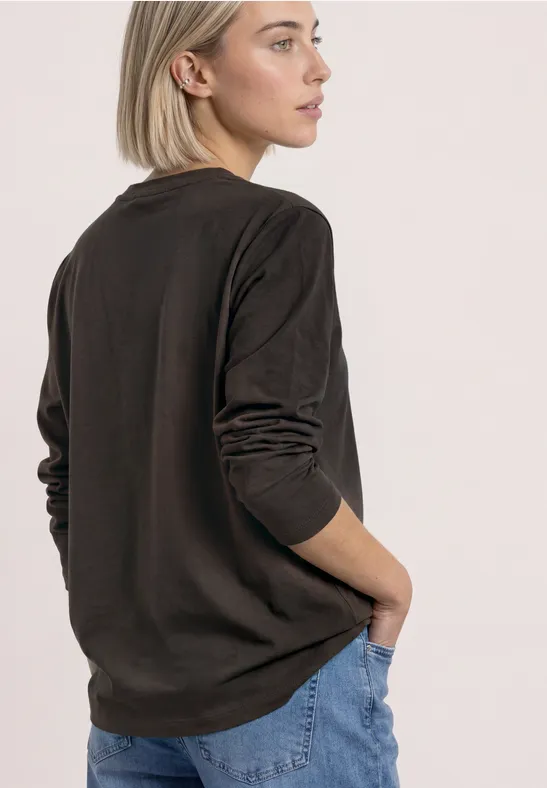 Eine Frau in braunem Langarmshirt und blauen Jeans steht seitlich abgewandt vor hellem Hintergrund.