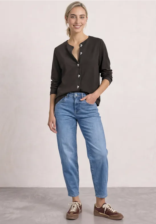 Eine lächelnde Frau trägt eine schwarze Bluse, blaue Jeans und zweifarbige Loafers.