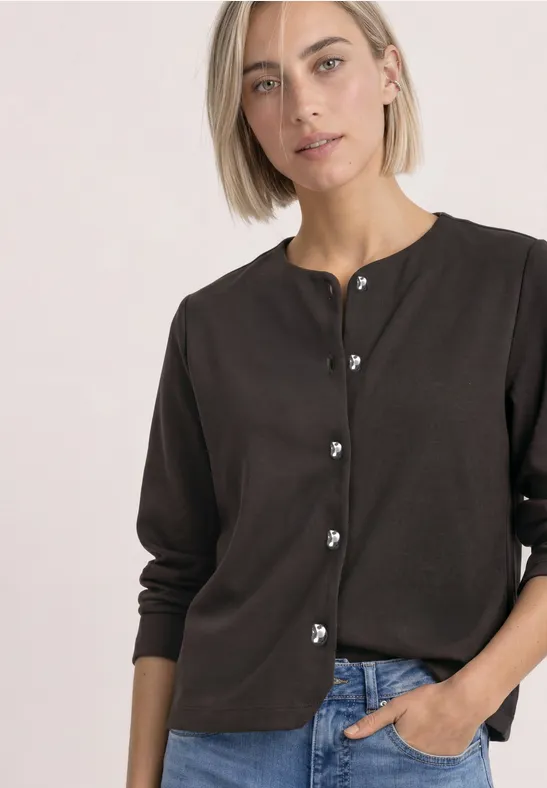 Frau mit kurzem blondem Haar trägt dunkelbraune Strickjacke mit Herzknöpfen, eine Hand in Jeans-Tasche.