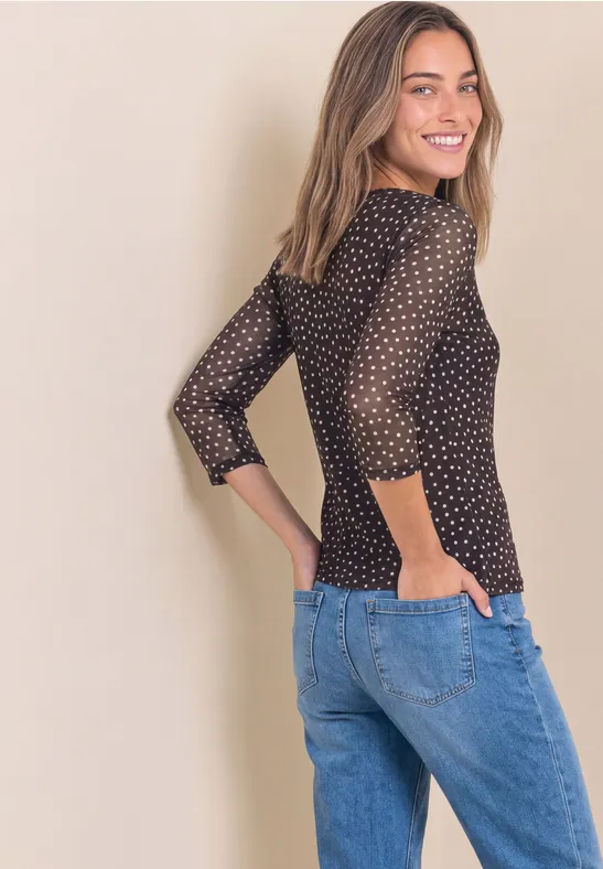Junge Frau im Profil trägt schwarzen Pullover mit weißen Punkten und blaue Jeans, lächelt über die Schulter.