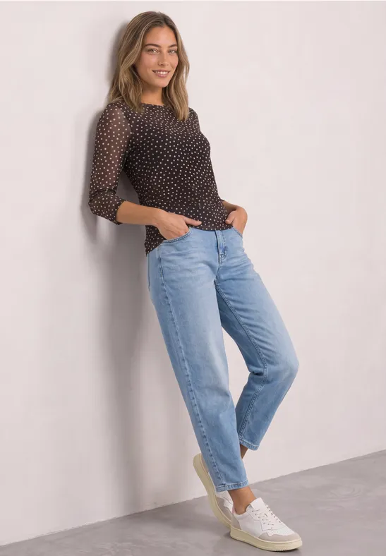 Frau lehnt an Wand, trägt gepunktetes Top, hellblaue Jeans und weiße Sneakers.