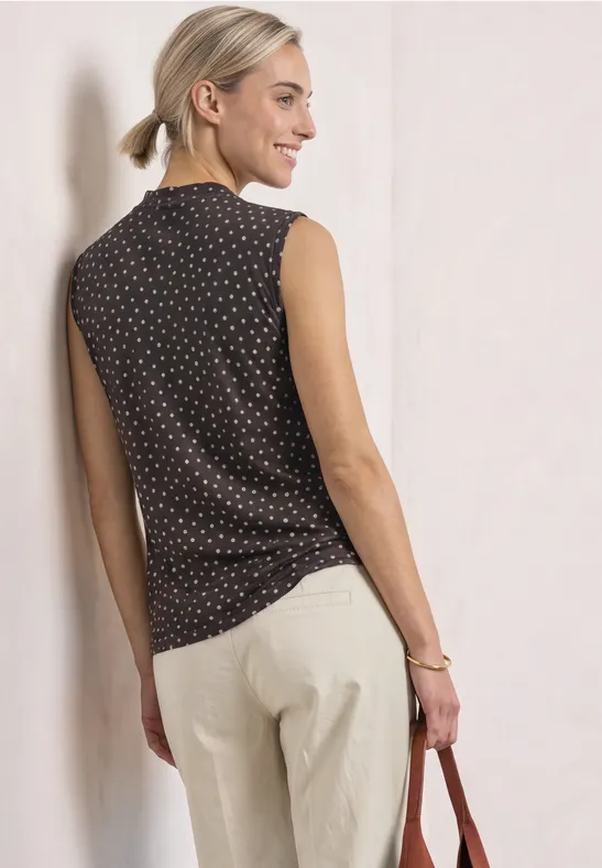 Frau in schwarzem ärmellosem Polka-Dot-Top und hellen Hosen hält eine Tasche und lächelt zur Seite.