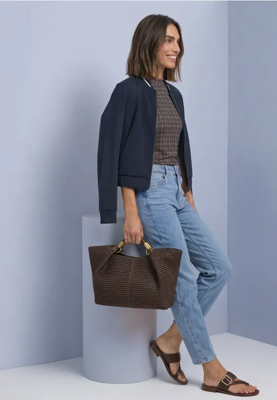 Femme en jean, top marron et veste bleue, souriante, appuyée devant un mur gris, tenant un sac marron.