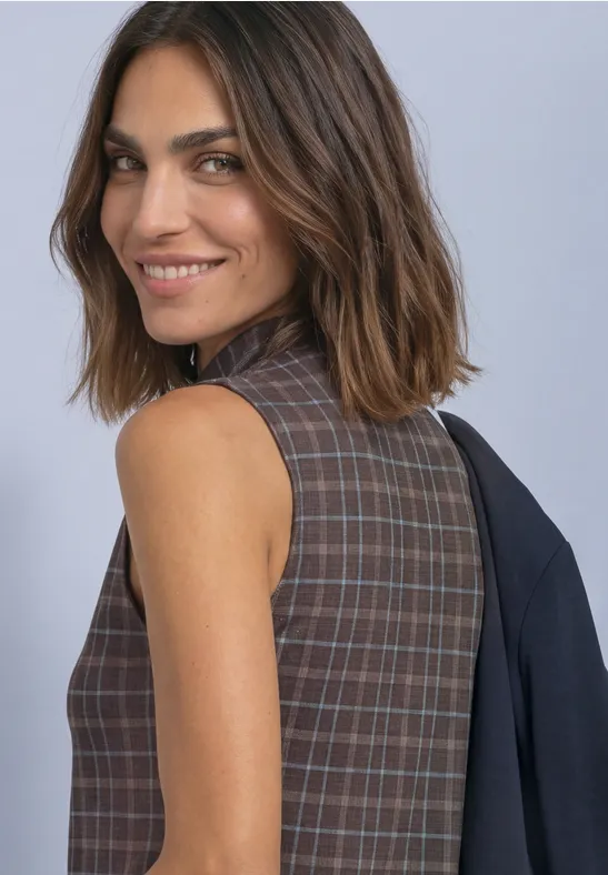 Une femme souriante porte une robe sans manches à carreaux, un blazer posé négligemment sur l’épaule, fond gris.