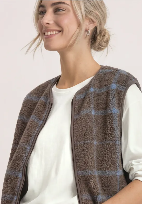 Eine lächelnde Frau trägt ein weißes Shirt und eine braune Fleeceweste mit dezentem hellblauem Karomuster.