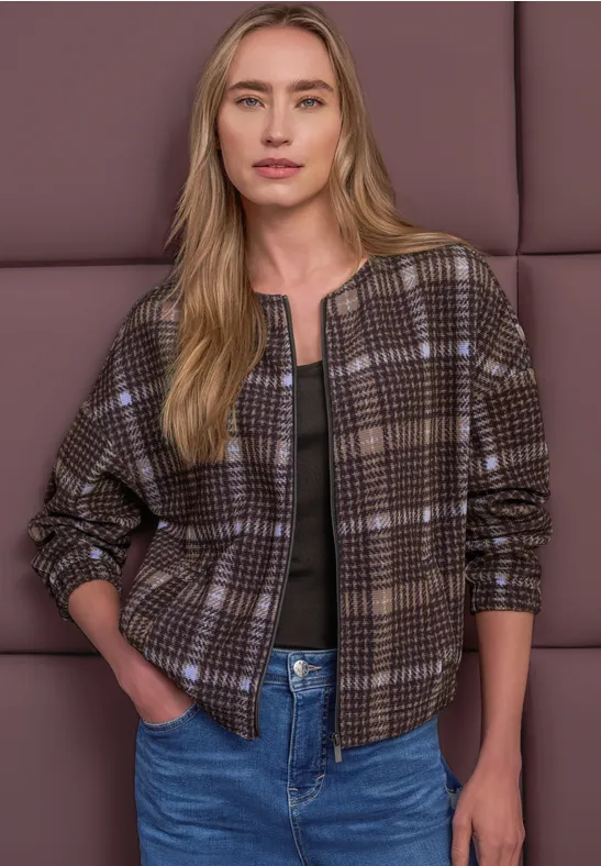 Blonde Frau in brauner, karierter Bomberjacke, schwarzem Shirt und Jeans, Hände in den Taschen.