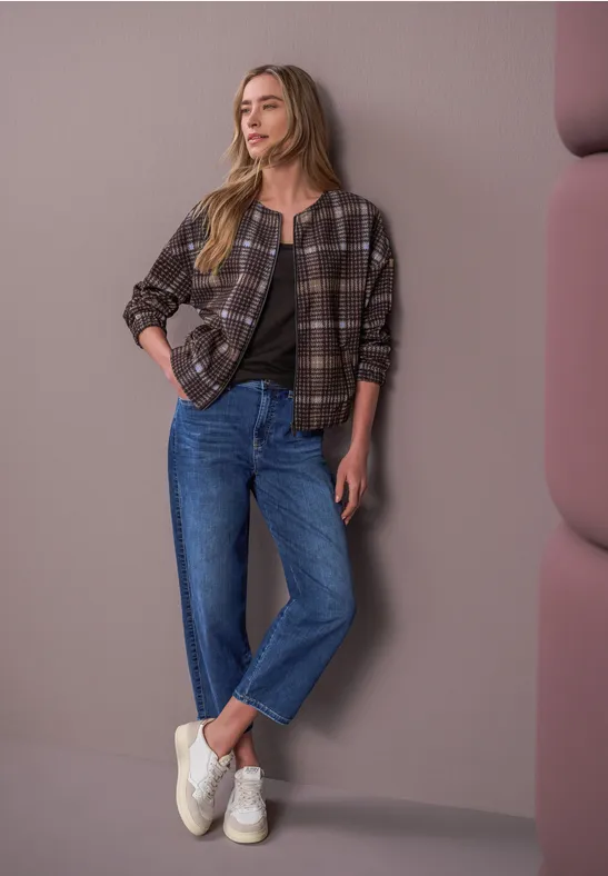 Eine Frau lehnt entspannt an einer Wand, trägt kariertes Hemd, blaue Jeans und weiße Sneakers.