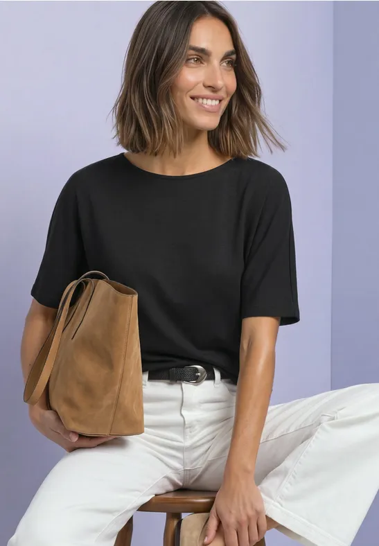 Eine lächelnde Frau sitzt auf einem Hocker, trägt schwarze Bluse, weiße Hose und hält eine braune Tasche.