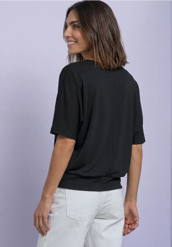 Eine Frau steht seitlich, trägt ein schwarzes T-Shirt und weiße Hose vor lila Hintergrund.